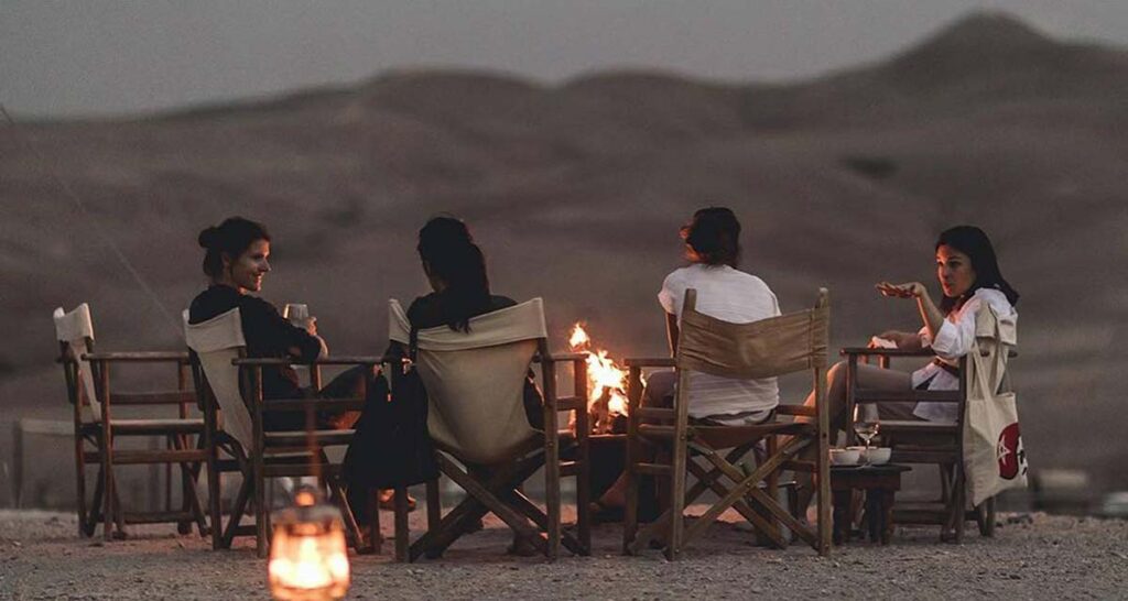 Four friends enjoy a candlelit dinner in the Agafay Desert, with rolling dunes under a twilight sky.